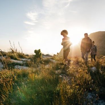 Group of people hiking through a grassy landscape at sunrise or sunset, with sunlight streaming behind them.